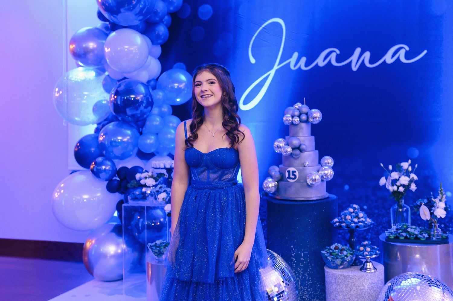Teen girl in blue dress poses at quinceañera party with balloon arch and name sign reading Juana.