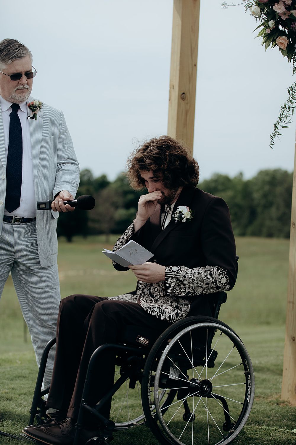 Emotional groom in wheelchair reading vows at outdoor wedding ceremony, wearing black floral suit with boutonniere.