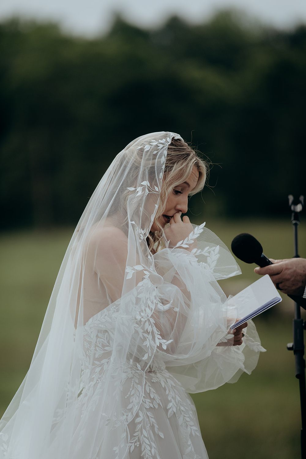 Emotional bride in lace veil reading wedding vows outdoors, holding a small book near a microphone.