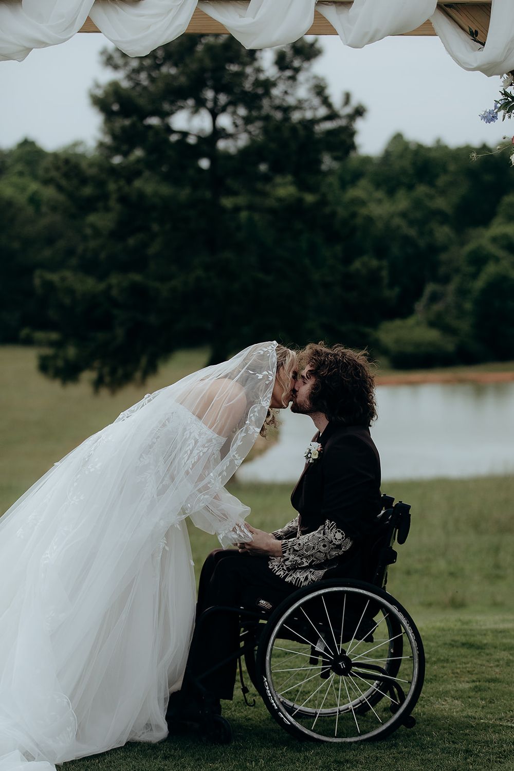 Bride in white gown kissing groom in wheelchair at outdoor wedding ceremony with lush green trees and pond backdrop.