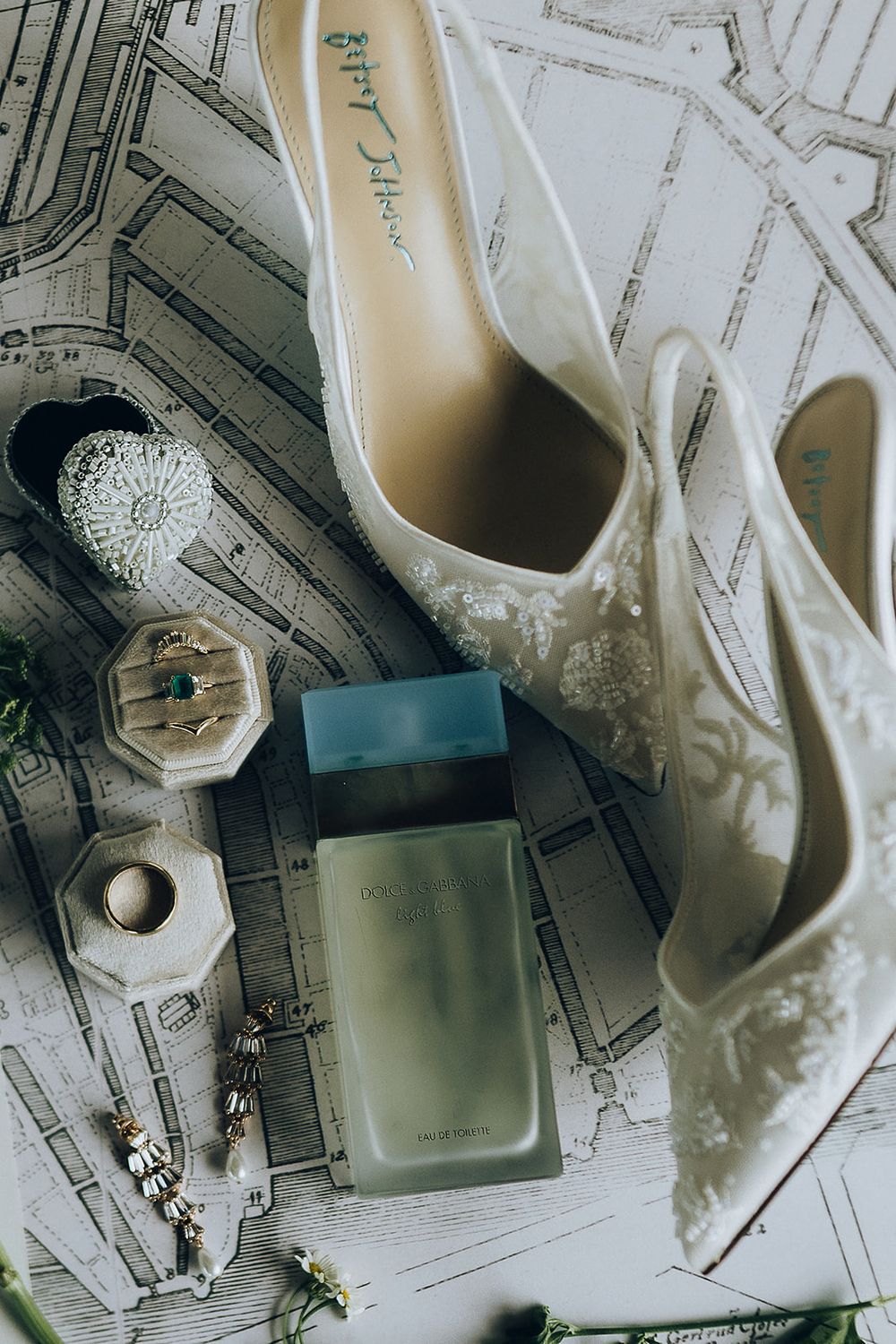 Elegant bridal flat lay featuring Dolce & Gabbana perfume, white lace heels, jewelry, and rings on a map background.