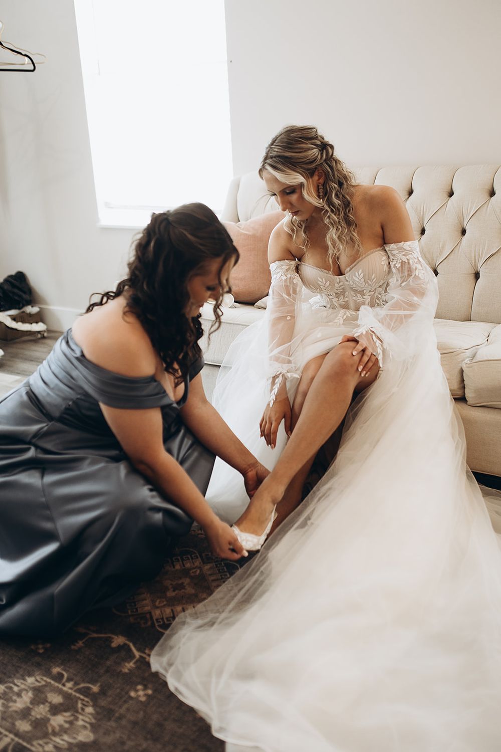 Bridesmaid in gray dress kneels to help bride put on wedding shoes before ceremony.