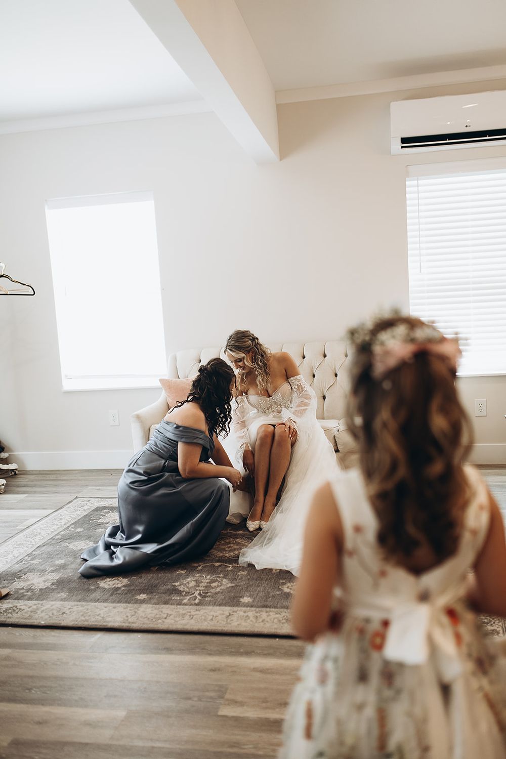 Bridesmaid assists bride with shoes as flower girl watches in bright wedding suite.