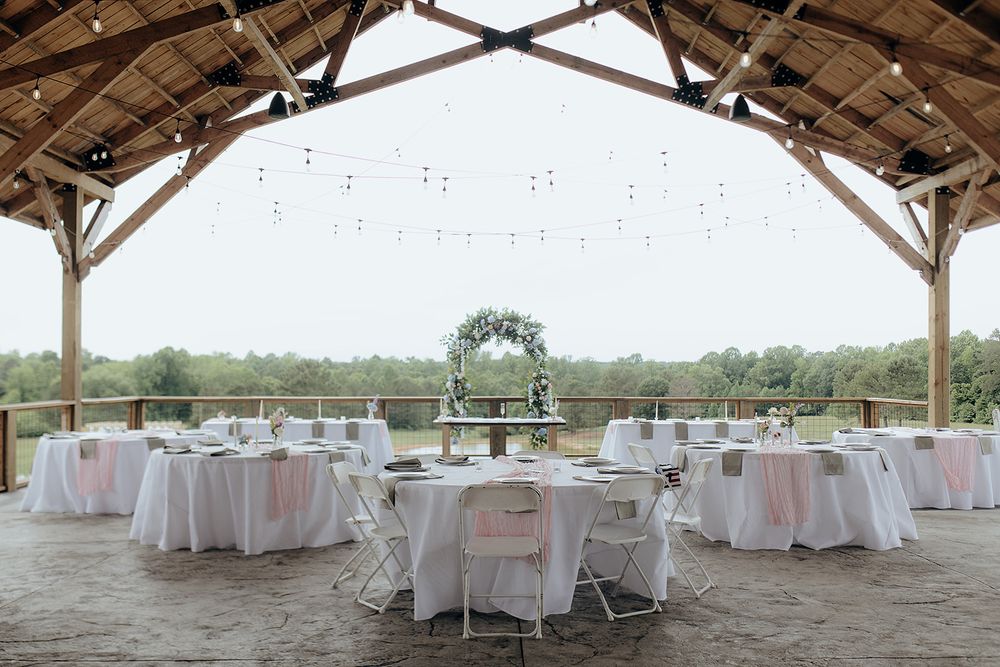 Elegant outdoor wedding reception under a wooden pavilion with white draped tables, string lights, and a floral arch backdrop.