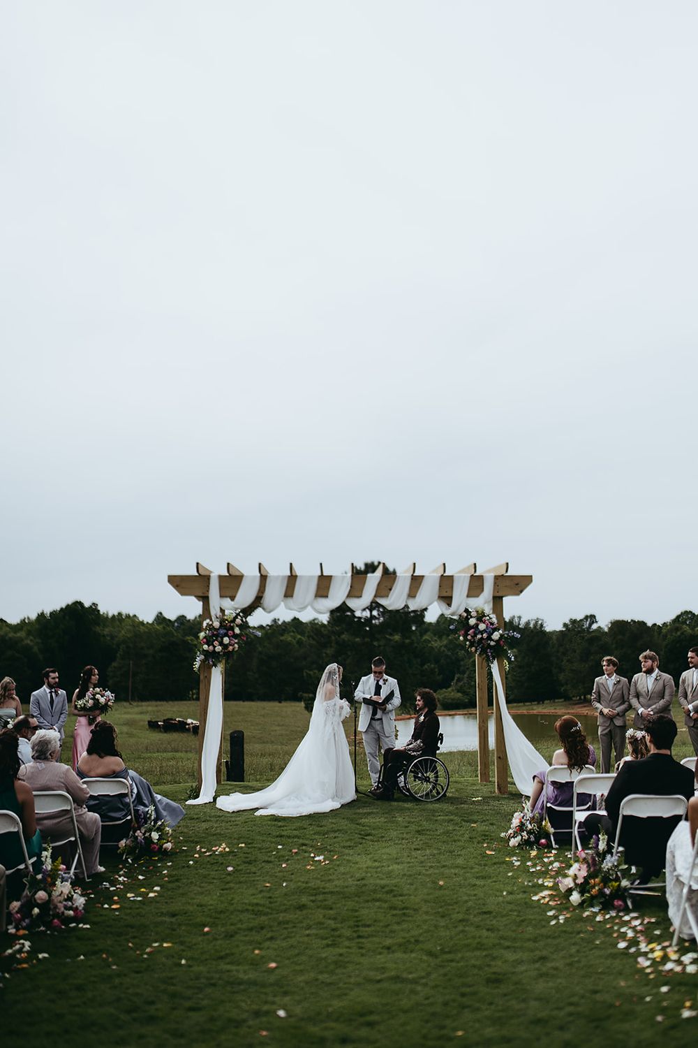 Outdoor wedding ceremony on green lawn with wooden arch, floral decor, bride in white gown, groom, and seated guests.