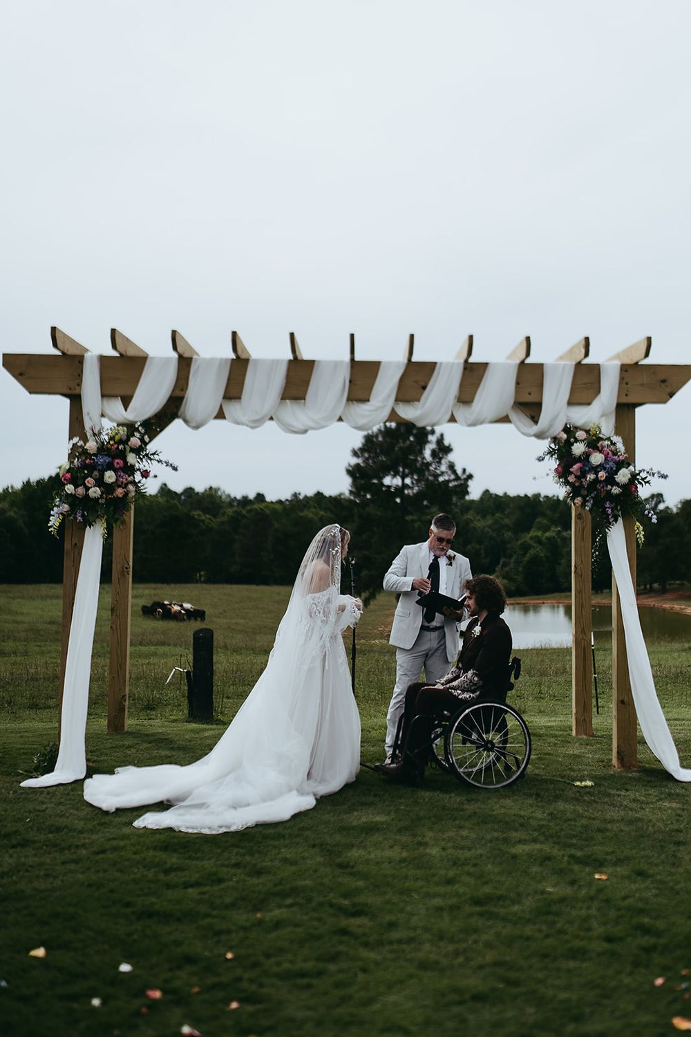Outdoor wedding ceremony under wooden pergola with white draping, bride in gown, groom in wheelchair, officiant reading vows.