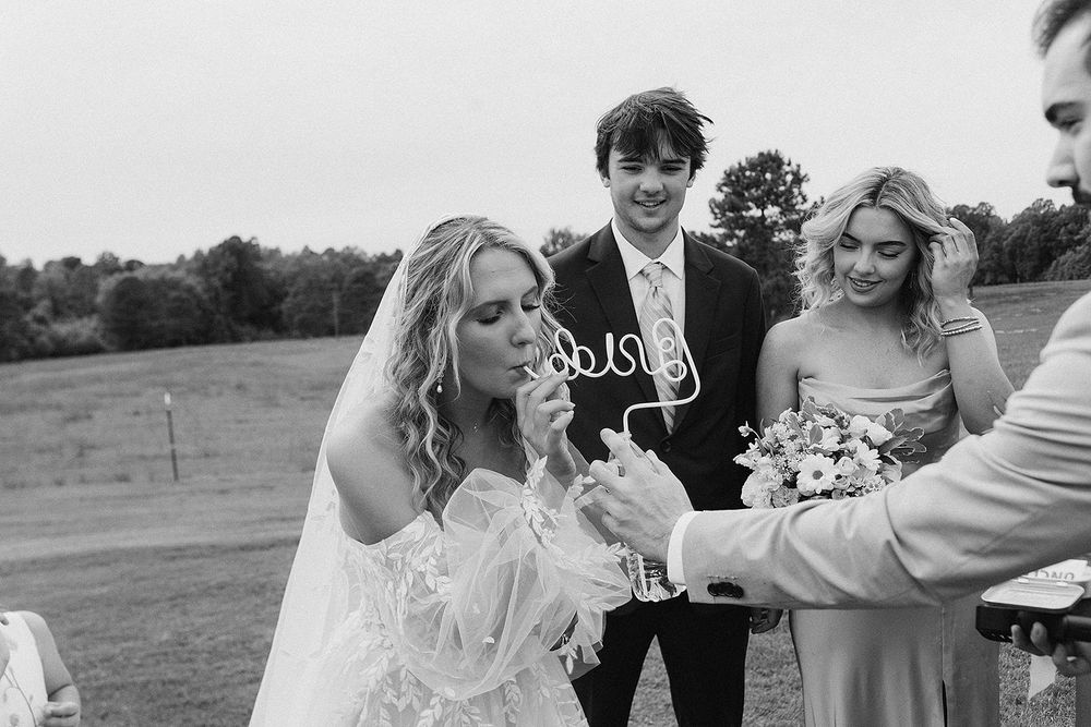 Black and white photo of smiling bride sipping drink with wedding party holding bouquet at outdoor ceremony.