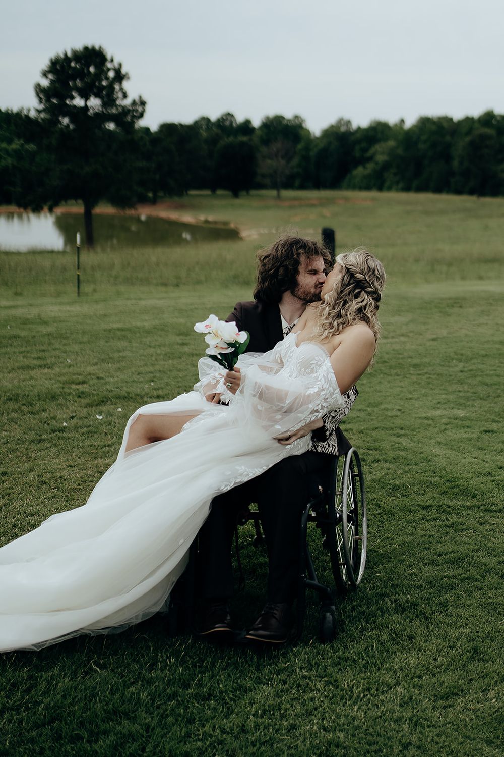 Bride in white lace gown sits on grooms lap in wheelchair sharing romantic kiss on lush green golf course.