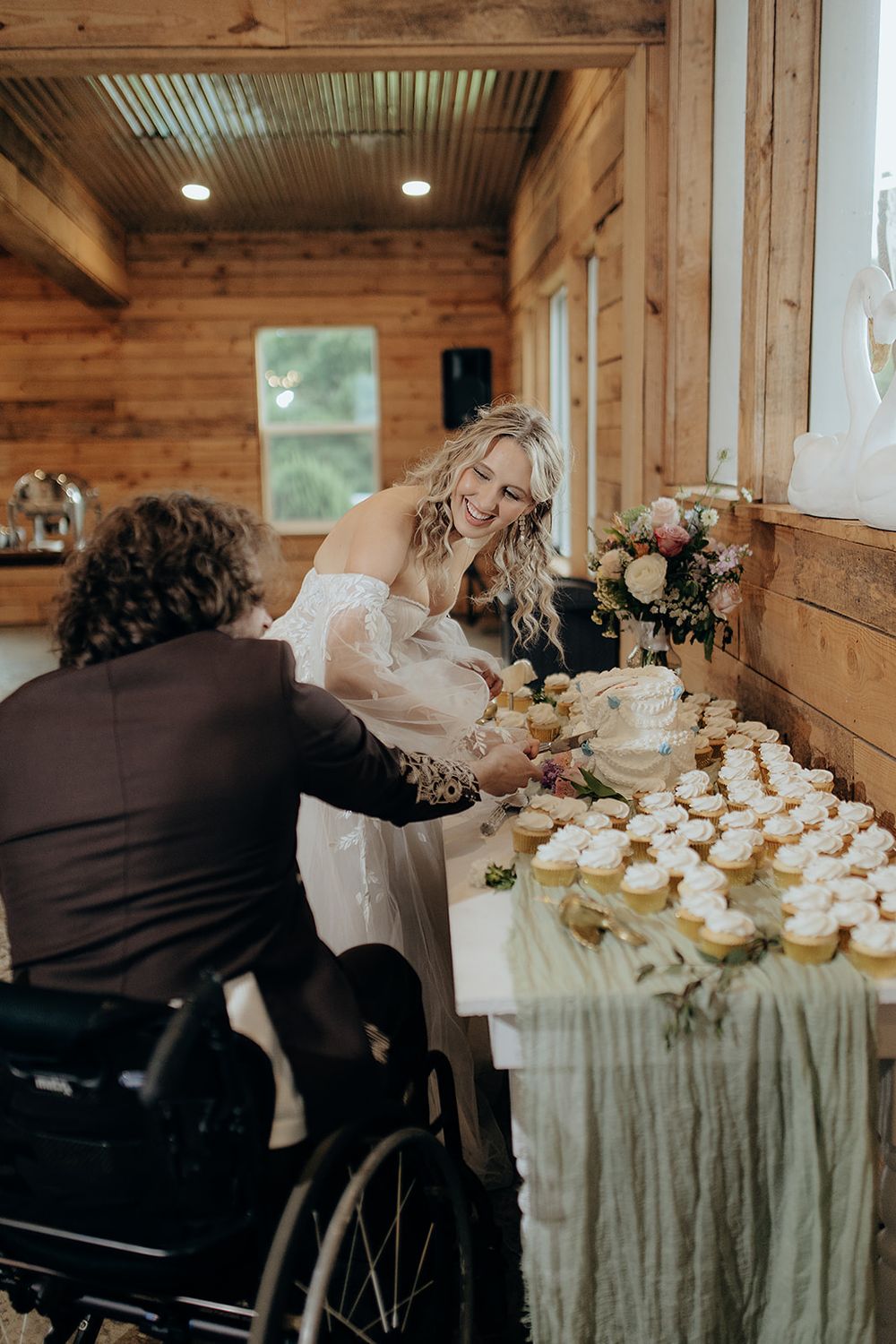 Bride smiling while cutting wedding cake with groom in wheelchair at rustic barn reception with cupcakes.