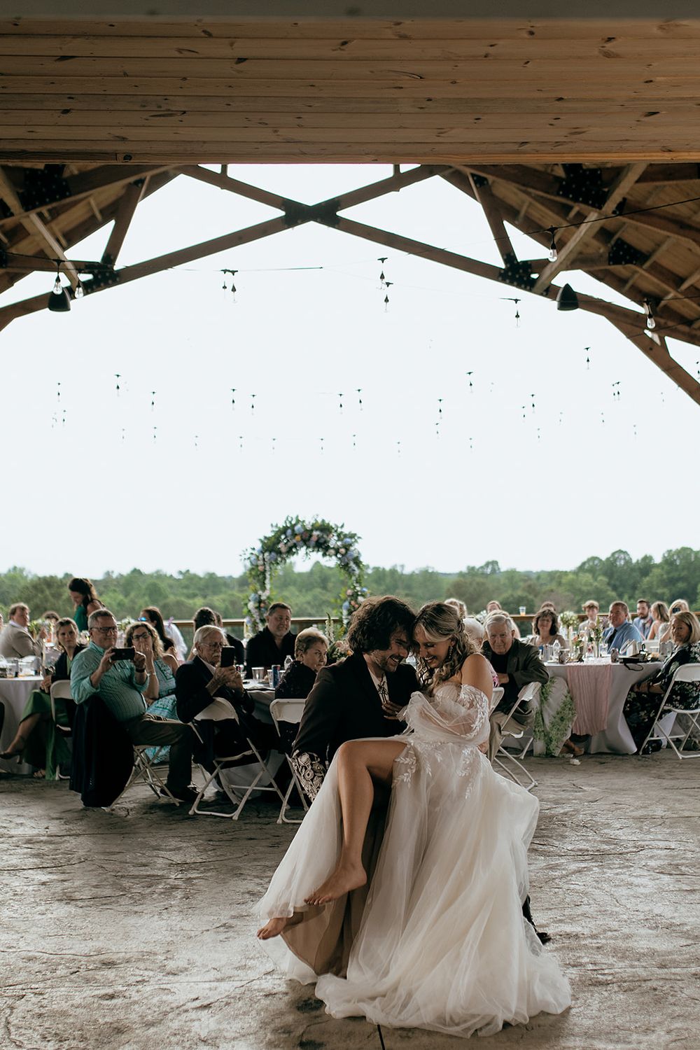 Bride and groom share first dance under rustic wooden pavilion with string lights at outdoor wedding reception.