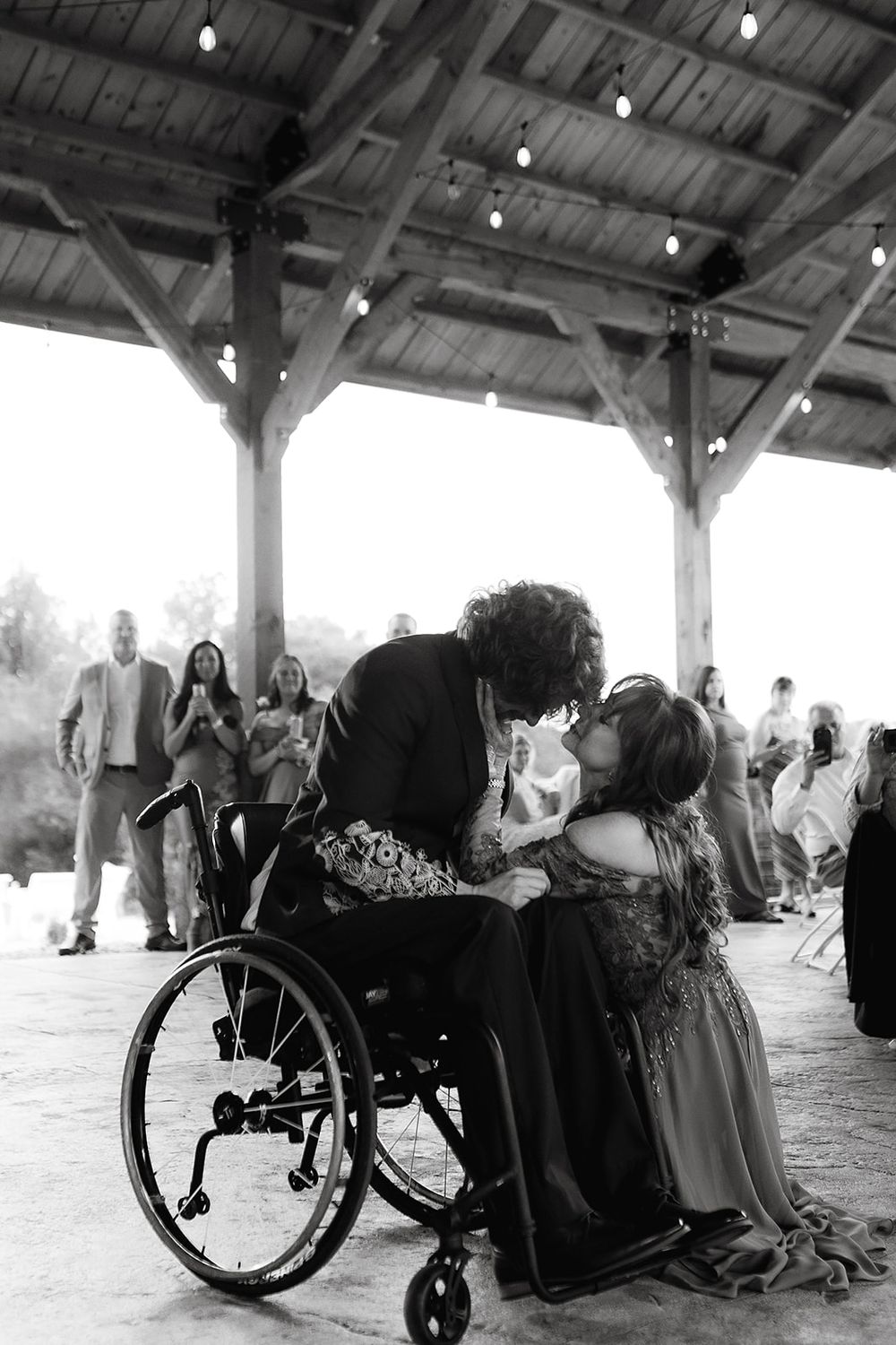 Black and white photo of a couple sharing a kiss, one seated in a wheelchair, at an outdoor wedding reception.