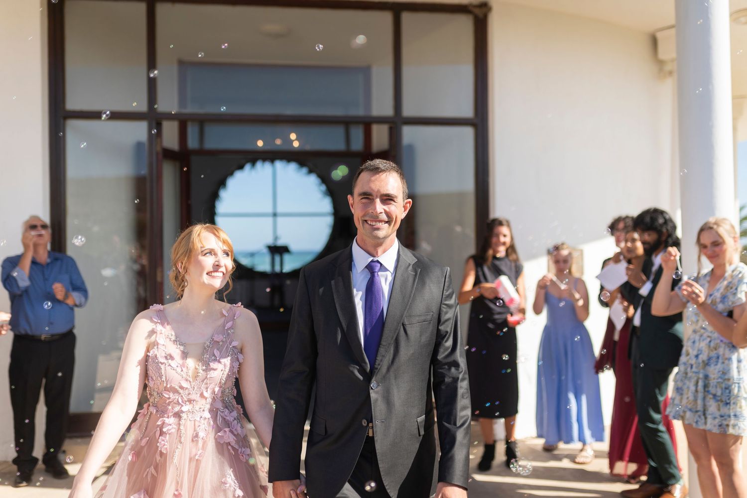 Newlywed couple walks out of wedding venue as guests celebrate with confetti in the background.