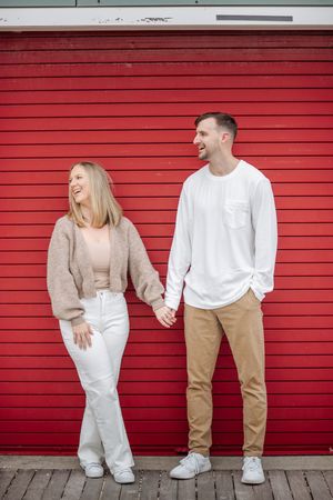 Happy couple holding hands and laughing in front of a bright red wall on a wooden boardwalk.
