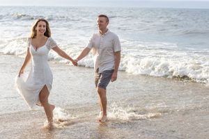 Smiling couple holding hands and walking barefoot along a sunny beach with ocean waves.