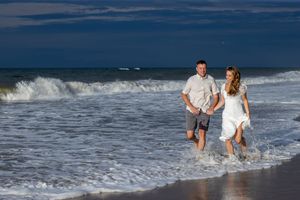 Couple running hand in hand through ocean waves on a beach under a dramatic blue sky.