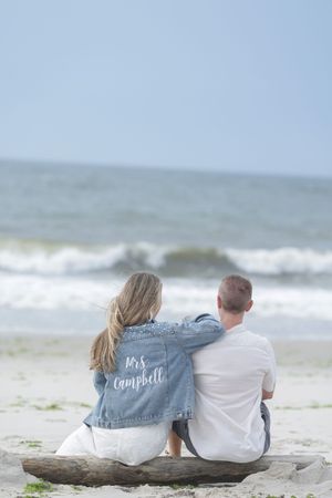 Couple sitting on driftwood facing the ocean, woman wearing a Mrs. Campbell denim jacket.
