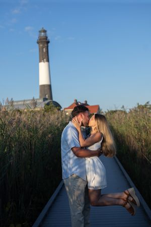 Couple kissing on a boardwalk with a tall lighthouse in the background.