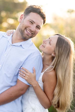 Smiling couple embracing outdoors in warm golden sunlight with engagement ring visible.