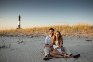 Couple sitting together on sandy beach with a lighthouse at sunset.