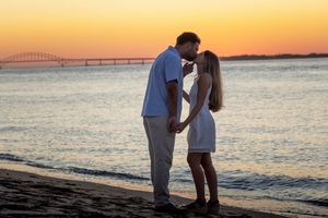 Couple kissing on a beach at sunset with a bridge visible over the water.