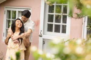 Man embracing woman smiling near a pink house with green foliage.