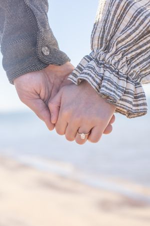 Close-up of couple holding hands on beach, woman wearing oval diamond engagement ring.