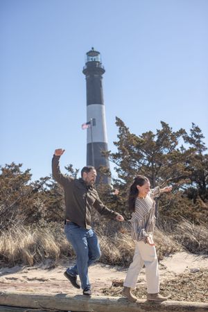 Joyful couple balancing on driftwood with iconic black and white lighthouse in background.