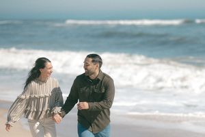 Happy couple laughing and holding hands while walking along a sunny ocean beach.
