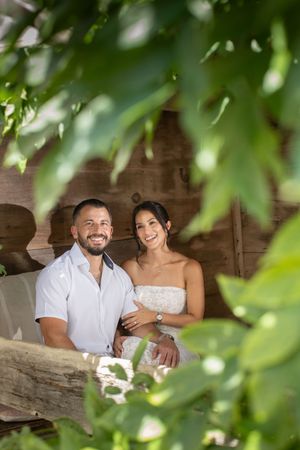 Smiling couple posing together framed by lush green leaves near a rustic wooden wall.