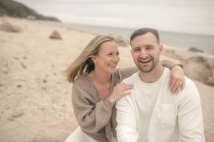Smiling couple sitting together on a sandy beach with ocean waves in the background.