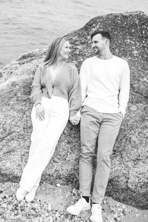 Black and white photo of a couple holding hands while leaning against large coastal rocks.