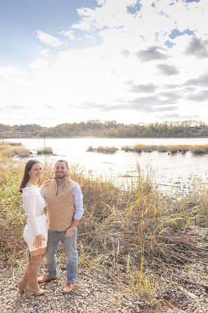 Couple posing together near a scenic lake surrounded by tall autumn grasses under a cloudy sky.