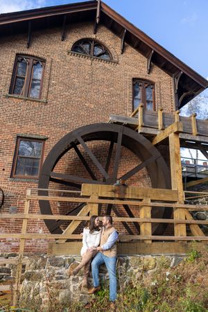 Couple sitting together in front of a historic brick mill building with a large wooden water wheel.