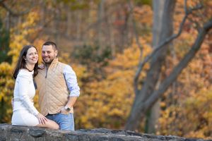 Smiling couple sitting on a stone wall surrounded by vibrant golden autumn foliage.