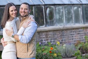 Happy couple embracing in front of a greenhouse with colorful flowers in a garden setting.
