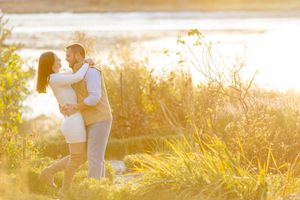 Romantic couple embracing and kissing in a sunlit field with golden autumn plants by a lake.