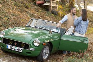 Couple standing beside a classic green MG convertible car surrounded by autumn foliage.