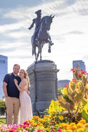 Couple embracing in front of an equestrian statue surrounded by colorful flowers in a city park.
