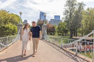 Couple walking hand in hand across a white ornamental bridge in Boston Public Garden.