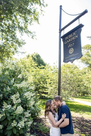 Couple sharing a kiss beneath Boston Public Garden 1837 sign surrounded by green trees and shrubs.