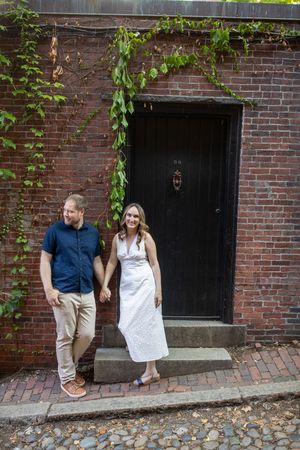 Couple holding hands and smiling outside a charming brick building with ivy-covered walls on cobblestone street.