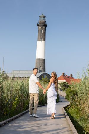 Couple holding hands on a boardwalk path with a black and white lighthouse towering behind them.