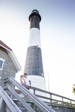 Romantic couple kissing on wooden stairs with a tall black and white striped lighthouse looming above.