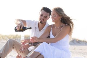 Happy couple sitting on beach sand while man pours champagne into glasses at sunset.