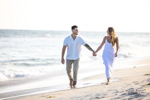 Couple holding hands and walking along a sandy beach shore with ocean waves behind them.
