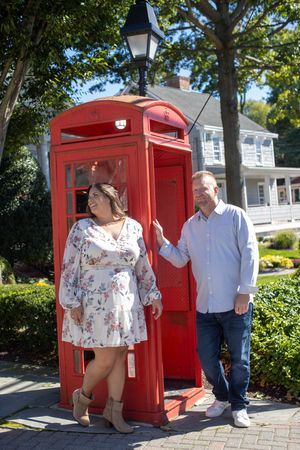 Smiling couple posing beside a classic red British phone booth in a charming town setting.