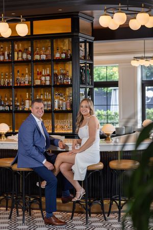 Elegant couple smiling at a stylish upscale bar with a well-stocked liquor shelf backdrop.