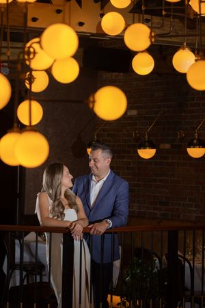 Couple shares romantic moment on balcony surrounded by glowing amber pendant lights in elegant brick-wall restaurant.