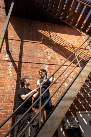 Couple laughing on a metal fire escape staircase by a brick wall.