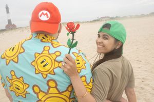 Woman smiling as man presents a Mario-themed metal rose on a sandy beach by a lighthouse.
